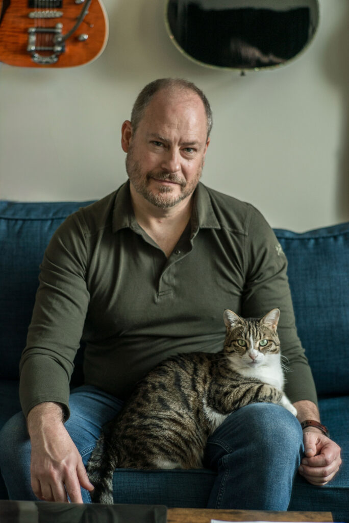 Sean Carberry seated on a couch with his cat Squeak in his lap. Guitars hang on the wall in the background.