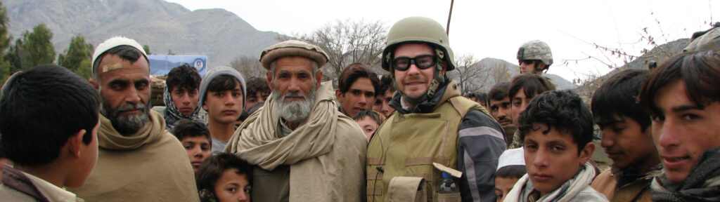 Sean Carberry standing with over a dozen men and boys in the street with a mountain range in the background. Sean is wearing a bullet-proof vest and a protective helment.
